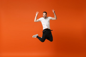 Fototapeta na wymiar Smiling young man in casual white t-shirt posing isolated on orange wall background in studio. People lifestyle concept. Mock up copy space. Having fun, fooling around, jumping, showing victory sign.