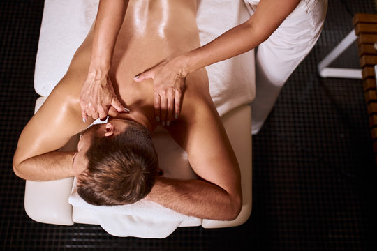 Young pleasant man having deep tissue massage on white soft towels, lying on black floor, gentle female hands massaging neck of male client, osteopathy treatment