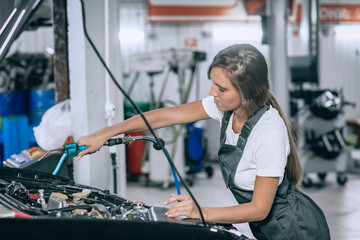 Caucasian womanl in a black jumpsuit and white t-shirt in overalls examining under hood of car in the repair garage