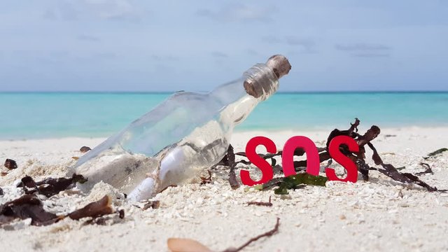A Glass Bottle Sealed With A Cork And A Rolled Up Paper Inside, Red colored wooden leters SOS and seaweed on white sand beach