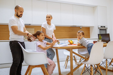 Obraz premium Caucasian family in white kitchen with modern interior taking meal. Children sit at the table, parents standing and look at them. Family, children, love, home concept