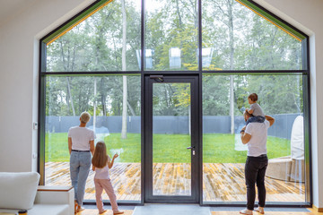 Obraz premium Back of Parents with children look at panoramic window together, relaxing together, spending time at home. Modern house interior