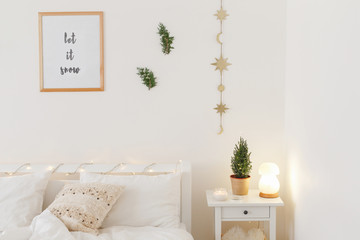 New year winter home interior decor. Christmas holiday decorations. White stylish cozy scandinavian bedroom: bed, bedside table, night lamp, little christmas tree in a pot, frame with text LET IT SNOW