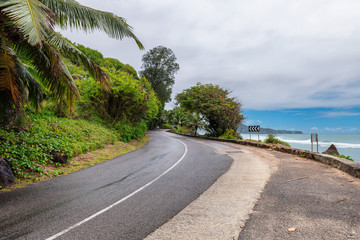 Road trip on island beach road with palm trees in ocean beach with dramatic sky on tropical island, Seychelles.