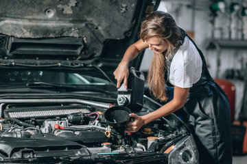 Naklejka na ściany i meble Beautiful Mechanic girl in a black jumpsuit and a white T-shirt changes the oil in a black car. car repair concept