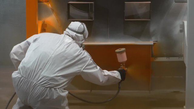 Man in protective suit squats down, holds spray gun in his hands, paints metal body of boiler in orange, factory, workshop, production of boiler rooms, manual labor, dangerous atmosphere for breathing