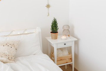 Little christmas tree in a pot and night salt lamp on bedside table. New year winter home interior decor. Christmas holiday decorations with led garland lights. White stylish cozy scandinavian bedroom