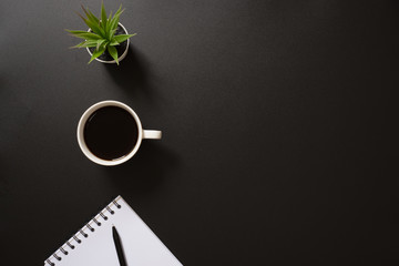 desk office top view with blank notepad, pen, coffee and plant on black background. flat lay with...