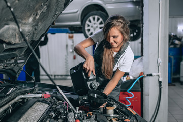 Obraz na płótnie Canvas Beautiful Mechanic girl in a black jumpsuit and a white T-shirt changes the oil in a black car. car repair concept