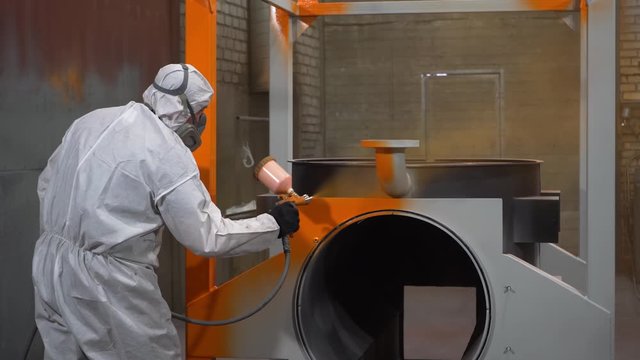 Factory worker paints frame of mobile boiler in orange using spray gun, paint, industrial painting, spacious workshop, man in chemical protection suit, respirator on face, hazardous work, production