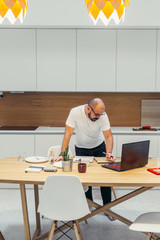 Busy caucasian bearded male working online from home, white colors interior. Modern kitchen. Using laptop every day