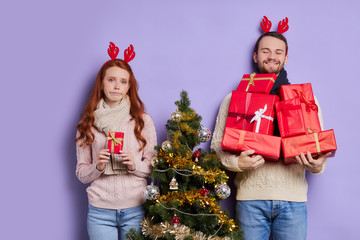 Tall handsome man satisfied by great number of gifts, holding presents with pleasure, thankful to friends, upset girl with small christmas gift in hands expressing disappointment