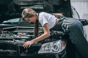 A brunette in a black jumpsuit and a white t-shirt near the open hood of black car. Young female in the garage is smiling at the camera and lowered gaze. car repair concept