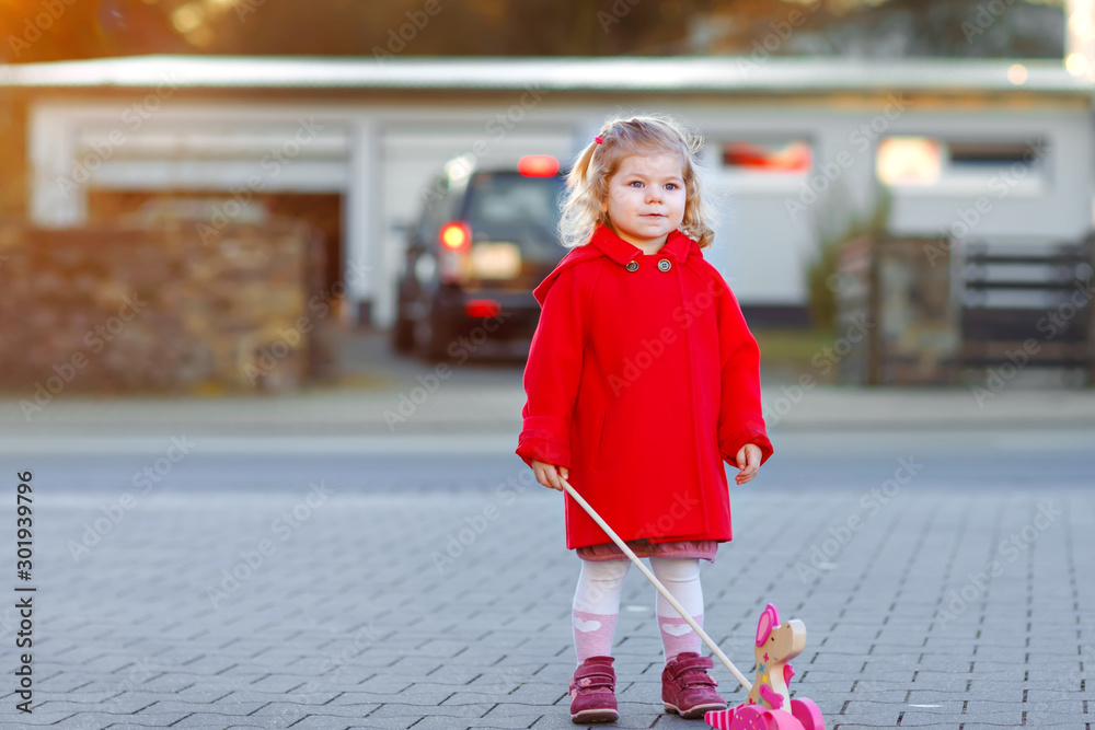 Canvas Prints Outdoor portrait of little cute toddler girl in red coat aon spring sunny day with push wooden toy. Healthy happy baby child walking in the city. Fashion stylish clothes for kids.