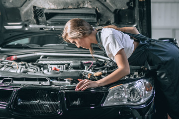Fototapeta na wymiar A brunette in a black jumpsuit and a white t-shirt near the open hood. Young female in the garage is smiling at the camera and lowered gaze. car repair concept