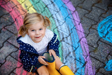 Happy little toddler girl in rubber boots with rainbow sun and clouds with rain painted with colorful chalks on ground or asphalt in summer. Cute child having fun. creative leisure - obrazy, fototapety, plakaty