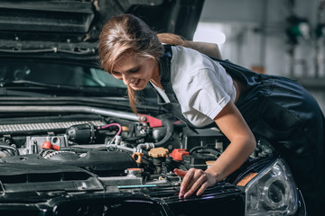 Naklejka na ściany i meble A brunette in a black jumpsuit and a white t-shirt near the open hood of black car. Young female in the garage is smiling at the camera and lowered gaze. car repair concept