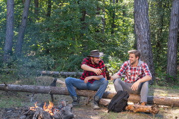 Young people enjoying picnic in park on summer day and drinking beer. Group of two male friends camping with marshmallows over a camp fire. Tourism concept.