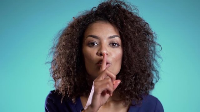 African american woman holding a finger on her lips over blue background. Gesture of shhh, secret, silence. Close up.