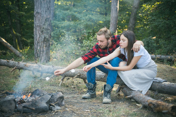 Pretty couple roast marshmallow candies on the campfire in forest. Tourists relaxing. Picnic time. Tourism concept. Young couple having picnic in woods. Camp fire in summer.