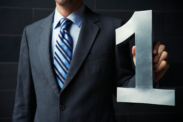 Unrecognizable businessman in a suit holding a large brushed steel number one against a dark background