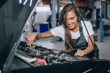 Fototapeta na wymiar A brunette in a black jumpsuit and a white t-shirt near the open hood of black car. Young female in the garage is smiling at the camera and lowered gaze. car repair concept