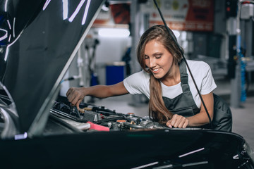 A brunette in a black jumpsuit and a white t-shirt near the open hood of black car. Young female in the garage is smiling at the camera and lowered gaze. car repair concept