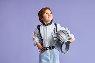 Smiling boy in white protective cosmonaut suit, holding helmet looking side, dreaming about flight to cosmos