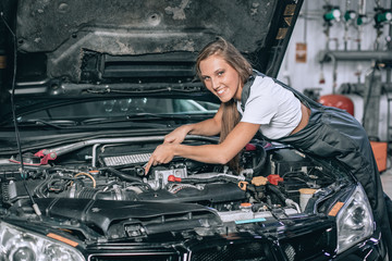 Fototapeta na wymiar A brunette in a black jumpsuit and a white t-shirt near the open hood of black car. Young female in the garage is smiling at the camera. car repair concept