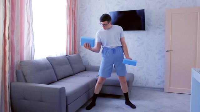 Funny nerd man in glasses and shorts is doing exercises for biceps with yoga blocks instead of dumbbells at home. Sport humor concept.