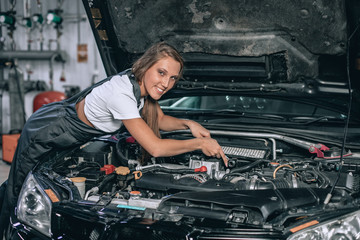 A brunette in a black jumpsuit and a white t-shirt near the open hood of black car. Young female in the garage is smiling at the camera. car repair concept