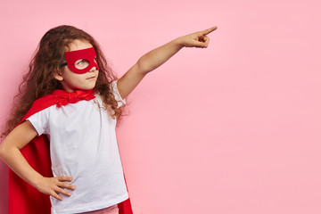 Attractive cute kid wearing red superhero suit and mask stand showing side isolated ove pink...