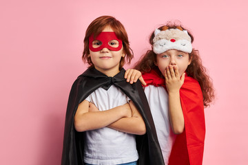 Two cute kids wearing hero cloaks and masks stand isolated over pink background, togetherness,...