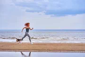 A red-haired middle-aged woman in sportswear runs along the sandy shore of a large river with her...