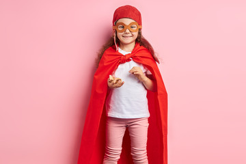 Fototapeta premium Portrait of caucasian kid girl wearing red cloak and helmet of pilot stand isolated over pink background. Girl hold wooden plane in hands