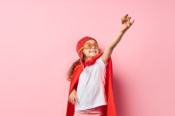 Fototapeta premium Słodkie dziecko dziewczyna ubrana w czerwony płaszcz i hełm pilota podniósł ręce do góry trzymając drewniany samolot zabawka, odizolowane na różowym tle