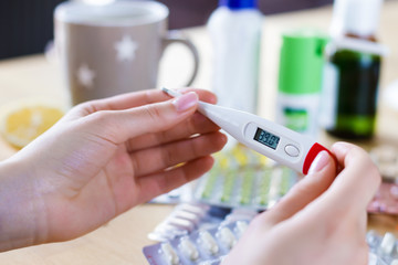 Closeup female hands are holding thermometer. Cup of tea, lemon, analgesic, antipyretic pills,...