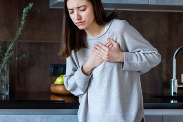 Young woman in grey clothes is holding hands on breast. Brunette girl is feeling bad. Sudden heart attack, myocardial infarction at home. Effect of stress and unhealthy lifestyle concept.