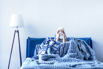 Sick exhausted girl in white warm hat wrapped herself in scarves and blankets is sitting in bed. Young woman with runny nose, fever and headache is treated at home. Winter cold and flu concept. - obrazy, fototapety, plakaty
