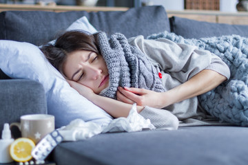 Sick exhausted girl in scarf is lying in bed wrapped in blanket. Young woman with fever and headache is measuring temperature with thermometer, treated at home. Winter cold and flu concept. - obrazy, fototapety, plakaty