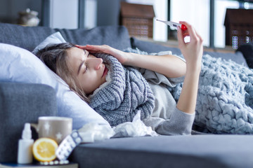 Sick exhausted girl in scarf is lying in bed wrapped in blanket. Young woman with fever and headache is measuring temperature with thermometer, treated at home. Winter cold and flu concept. - obrazy, fototapety, plakaty