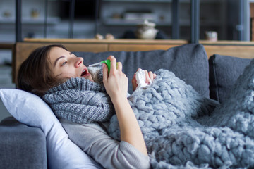Sick exhausted girl in scarf is lying in bed sofa wrapped in grey blanket. Young woman with runny nose, fever and sore throat is using .healing spray. Home treatment. Winter cold and flu concept. - obrazy, fototapety, plakaty