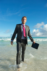 Exhausted young businessman in a full soaking wet suit holding his tablet computer walks from the crashing surf onto a tropical beach