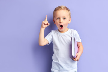 Caucasian child show bright emotions standing over purple background, holding purple notebook. With opened mouth
