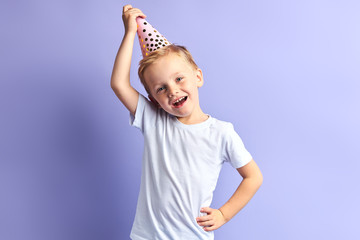 Portrait of happy little boy wearing birthday cap on head, isolated over purple background
