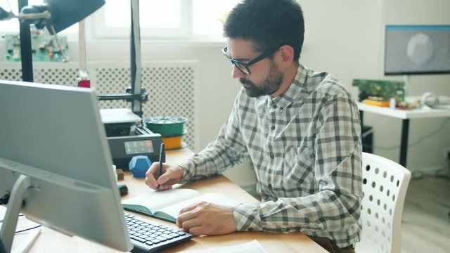 Slow motion of serious young man in glasses using computer and making 3d printer element in modern office. Technology and contemporary business concept.