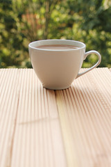 white cup with coffee on a light wooden table against a green summer garden, close-up, copy space