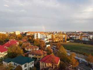 a quarter of a rainbow over a city in Romania on a cloudy autumn day. many houses to the left of the frame and an old stadium to the right. Background wallpaper image