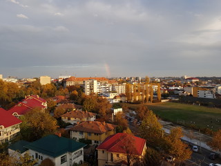 a quarter of a rainbow over a city in Romania on a cloudy autumn day. many houses to the left of the frame and an old stadium to the right. Background wallpaper image