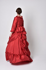 full length portrait of a brunette girl wearing a red silk victorian gown. Standing pose, with back to the camera,  on a white studio background.